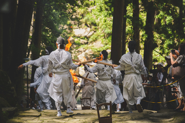The initial twelve, carrying the holy flame, make their way one last time down the mountain where the 50 kilogram-sized torches await to be lit. Wooden pails of water are placed every few steps apart in case of emergency. It is common to see volunteers take a scoop of water in their mouth, and spray it at the flames. After, the stone steps are a mix of damp from the falls, the buckets, and blackened by the ash.
