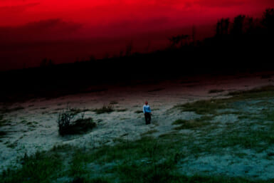 A man standing on sandy ground dotted with green brush gazes up into a red, cloudy sky. A dark tree-covered horizon looms behind him, and he appears small in the open landscape.