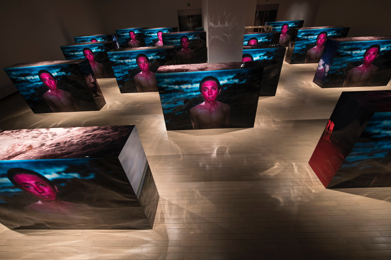 Photographic prints are wrapped around all four sides and the tops of some 15 boxes, which each stand about three meters high. The images on the fronts display the same shirtless, red-faced man, who stares straight ahead with a dark, rocky shore behind him. The images on the sides and tops of the boxes are all different: some show people, others ocean landscapes. 